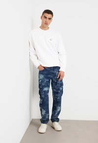 Valge sviiter, millel on rindkeres väike logo, koos siniste cargo-teksadega, millel on tekstureeritud ja kulunud muster. Valged spordijalatsid täiendavad riietust.