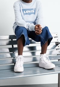 Personne portant un sweat-shirt blanc Levi's, un jean bleu court et des baskets blanches haute assise sur un banc en métal les mains jointes.