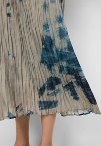 Plissé beige midi-rok met onregelmatige blauwe tie-dye patronen, gedragen door een persoon tegen een effen achtergrond.
