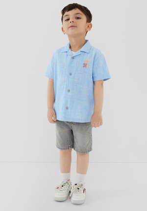 Młody chłopiec stoi, ubrany w jasnoniebieską koszulę z krótkim rękawem zapinaną na guziki, szare szorty, białe skarpetki i białe trampki na jednolitym tle.