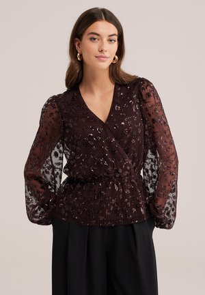 Femme aux longs cheveux bruns portant des boucles d'oreilles créoles dorées et un chemisier noir transparent à manches longues avec des motifs floraux pailletés et un pantalon noir.