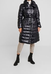 Abrigo acolchado negro con cinturón, cuello alto y acabado brillante. Presenta un patrón acolchado y botones en la parte delantera. Estilizado con pantalones de cuadros y botas.