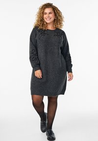 Tmavošedé pletené šaty s dlhými rukávmi, zdobené vyšívanými čiernymi kvetinovými vzormi na pleciach. V kombinácii s čiernou obuvou.