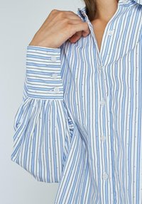Blauw-wit gestreepte blouse met knoopsluiting aan de voorkant, opbollende mouwen en kleine decoratieve details. Zachte stof met een lichte textuur.