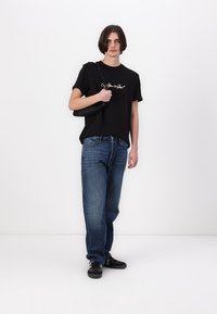Camiseta de algodón negra con un diseño de texto dorado, combinada con jeans de mezclilla azul. El modelo lleva una bolsa negra y calza zapatos negros.