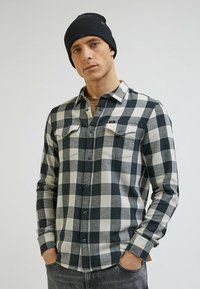 Czarna i biała w kratę koszula flanelowa z guzikiem z przodu oraz dwiema kieszeniami na klatce piersiowej. Noszona z ciemnym dzianinowym czapką. Szare dżinsy.