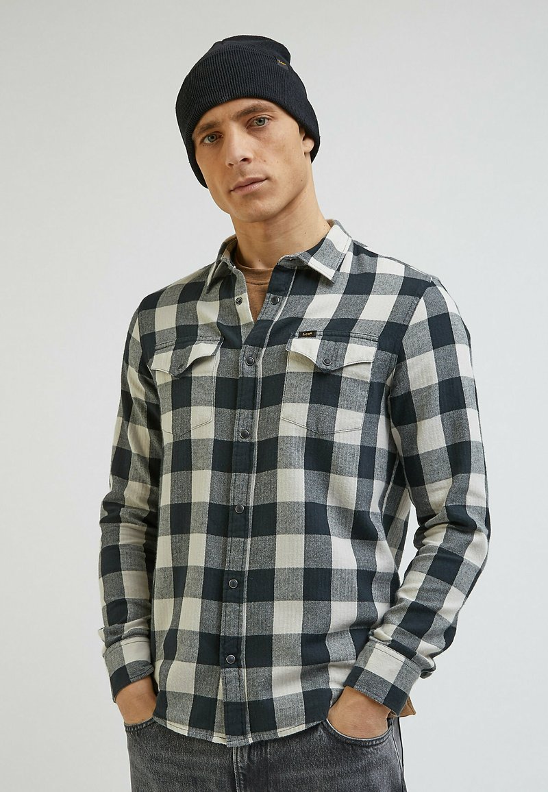 Czarna i biała w kratę koszula flanelowa z guzikiem z przodu oraz dwiema kieszeniami na klatce piersiowej. Noszona z ciemnym dzianinowym czapką. Szare dżinsy.