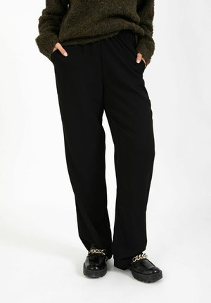 Személy laza, fekete nadrágot és fekete, aranyláncos loafereket visel, kezét a zsebében tartja, és sötétzöld, texturált pulóvert visel.