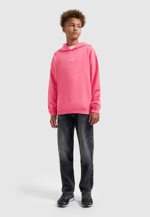 Tiener in een felroze hoodie, zwarte spijkerbroek en zwarte sneakers, staand tegen een effen lichte achtergrond, kijkend naar de zijkant.