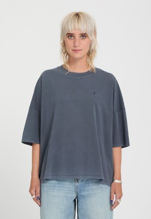 Personne aux cheveux blonds courts portant un t-shirt ample gris foncé et un jean bleu clair, debout devant un fond blanc uni.