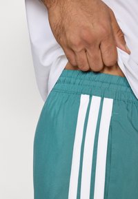 Türkiz atlétikai rövidnadrág fehér háromcsíkos részlettel, elasztikus derékrésszel és könnyű anyagból, amely részben fehér felső takar.