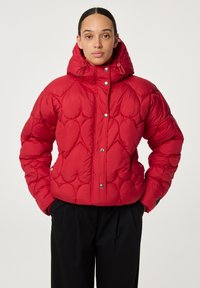 Vrouw in een rode gewatteerde pufferjack met capuchon en drukknoopjes, gecombineerd met zwarte broeken, staande met handen in de zakken.