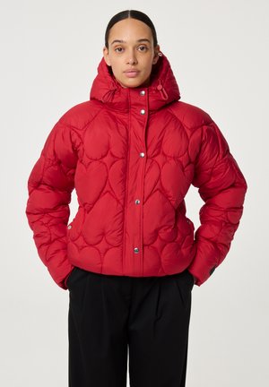 Vrouw in een rode gewatteerde pufferjack met capuchon en drukknoopjes, gecombineerd met zwarte broeken, staande met handen in de zakken.