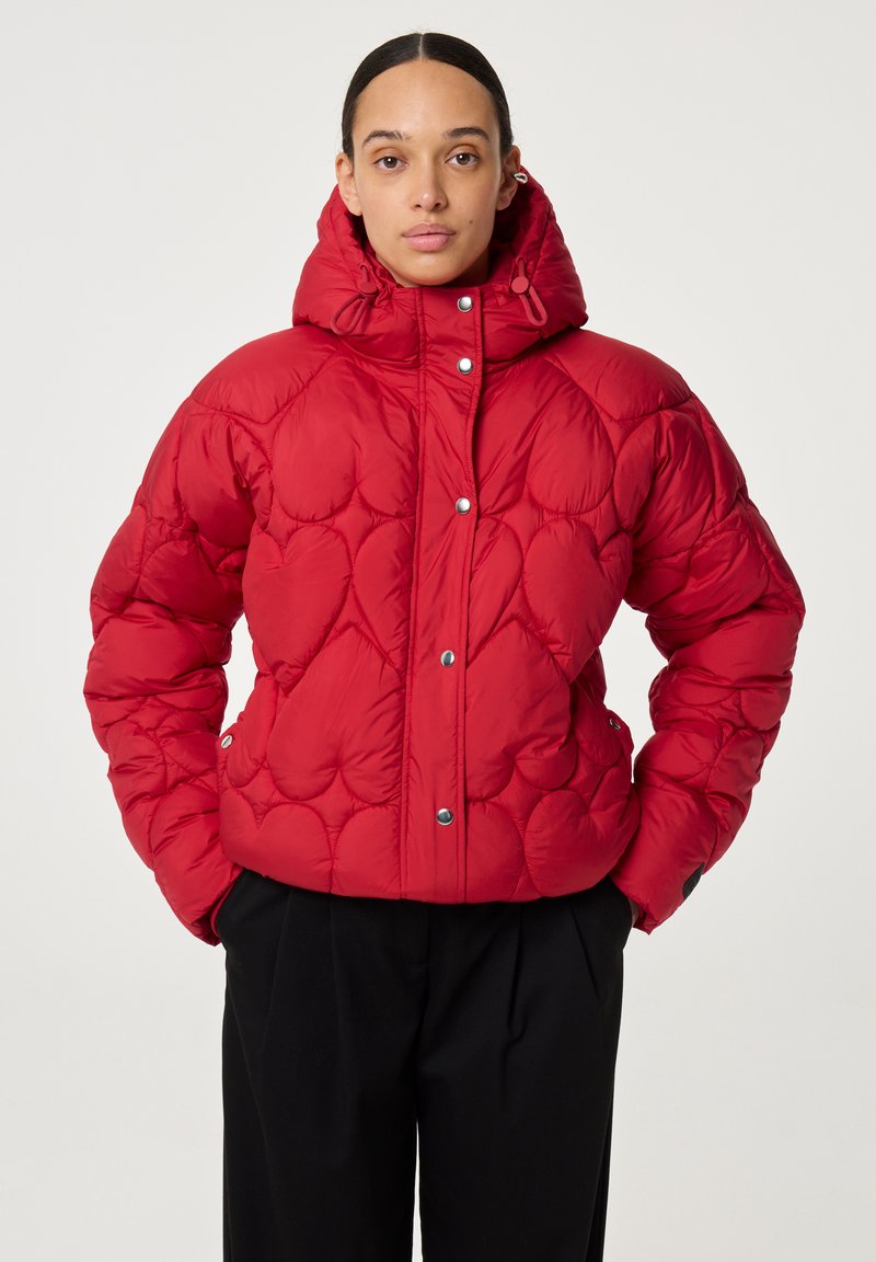 Vrouw in een rode gewatteerde pufferjack met capuchon en drukknoopjes, gecombineerd met zwarte broeken, staande met handen in de zakken.