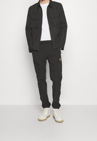 Black lightweight jacket with two front pockets, paired with black cargo pants featuring side pockets and a logo. White sneakers with beige sole.