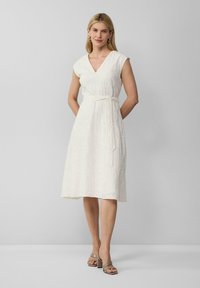 Robe blanche jusqu'au genou avec un décolleté en V, des manches courtes et une taille ceinturée. Tissu texturé avec des motifs subtils. Portée avec des escarpins beiges.