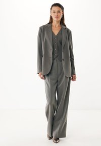 Traje gris a medida con un blazer estructurado, chaleco entallado con botones y pantalones de pierna ancha. Tela suave con una sutil textura y brillo.