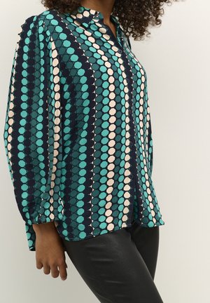 Femme portant un chemisier à manches longues à motif hexagonal turquoise, marine et beige avec un pantalon noir sur un fond uni.