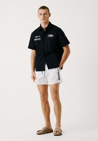 Camisa negra de botones de manga corta con logotipos, pantalones cortos de baño blancos con acentos azules y sandalias de lona marrón claro. Look sencillo y casual.