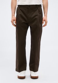 Άνδρας που φοράει σκούρα καφέ παντελόνια με ίσια γραμμή, λευκά αθλητικά παπούτσια με καφέ σόλες και ένα λευκό πουκάμισο κουμπωμένο μέσα, φαίνεται από τη μέση έως τα πόδια.