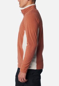 Seitenansicht einer Person, die eine rostfarbene Fleecejacke mit cremefarbenen Seiteneinsätzen und schwarze Hose trägt, vor einem weißen Hintergrund steht.