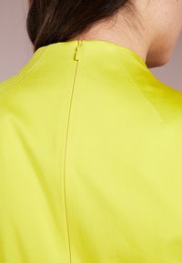 Žlutý textilní top s vysokým výstřihem, jemnými švy a zipovým zapínáním na zadní straně. Hladká textura a dobře padnoucí střih.