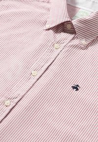 Rood-wit gestreepte blouse gemaakt van katoen. Heeft een kraag, knoopsluitingen en een marineblauw geborduurd logo op de borst.