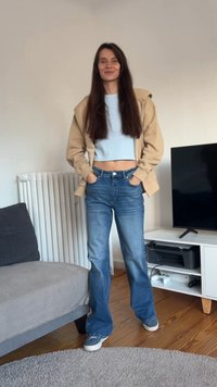 Beige hoodie, light blue crop top, and high-waisted blue jeans. Model stands on wooden floor, with gray couch and a TV in the background.
