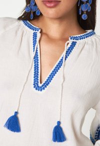 Blusa bianca con ricami floreali blu lungo il collo e nappine alle estremità del cordoncino. Il tessuto testurizzato conferisce profondità.