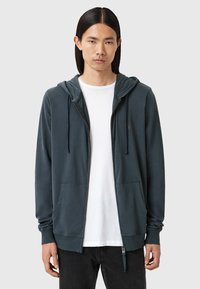 Felpa con zip di colore teal scuro, realizzata in morbido tessuto, con cappuccio, due tasche frontali e una maglietta bianca sotto. I jeans neri completano il look.