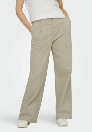 Femme portant un pantalon ample beige avec des poches latérales et des baskets blanches, debout les mains dans les poches devant un fond uni.