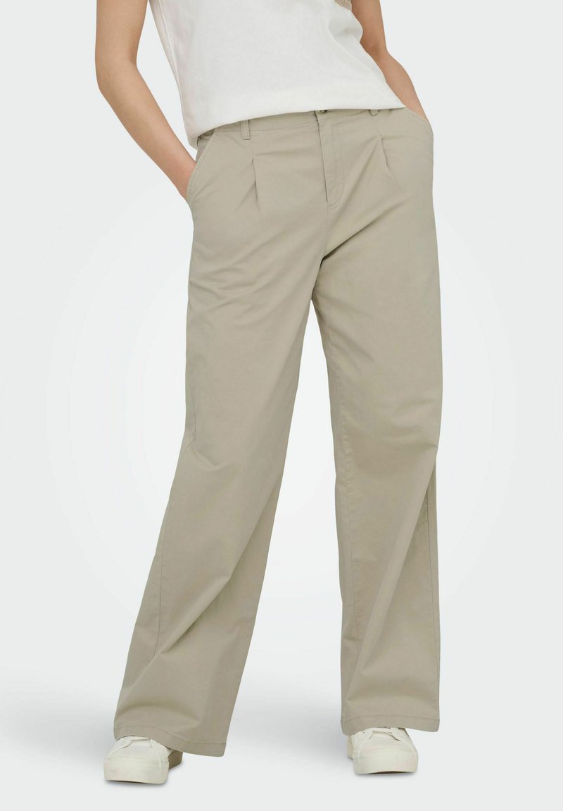 Femme portant un pantalon ample beige avec des poches latérales et des baskets blanches, debout les mains dans les poches devant un fond uni.