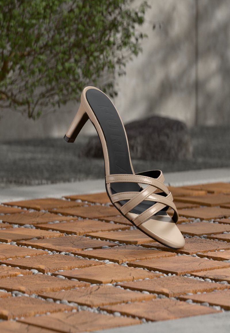 Sandale beige à talons hauts avec des lanières croisées flottant au-dessus d’un sol en bois carrelé, avec de la verdure et un mur gris en arrière-plan.