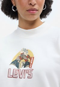Balta sporta krekla ar grafisku printu, kurā attēloti divi cilvēki plānotā un džinsa apģērbā. Levi's logo sarkanā krāsā ar punktveida akcentiem. Ribota apkakle.