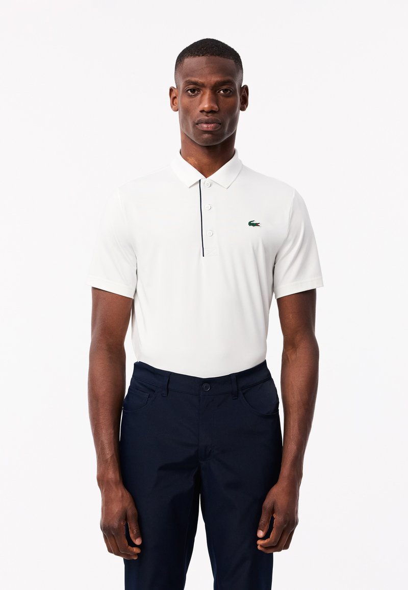 Polo shirt blanc à col court, arborant un logo Lacoste vert, assorti à un pantalon marine. Tissu lisse, coupe ajustée.
