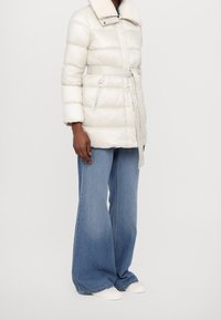 White puffer coat with a belted waist, metallic accents, and a structured collar. Paired with wide-leg blue jeans and white shoes.