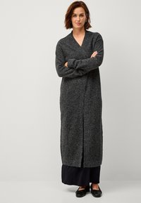 Langer grauer Cardigan aus weichem Material mit V-Ausschnitt, offener Vorderseite und seitlichen Schlitzern, kombiniert mit einem dunklen Rock und schwarzen Schuhen.