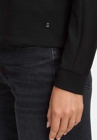 Czarny top z długim rękawem o gładkiej fakturze, z metalicznym przyciskiem w kolorze srebrnym blisko dolnej krawędzi. W połączeniu z ciemnymi dżinsami.