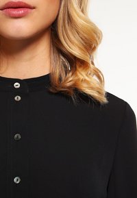 Blusa nera con colletto, caratterizzata da una chiusura frontale con bottoni in tono argento. Tessuto liscio con una leggera lucentezza e morbida consistenza.