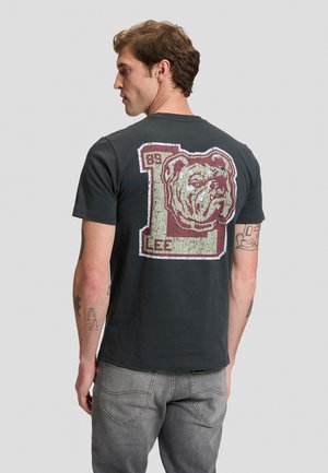 Homme aux cheveux bruns portant un t-shirt noir avec un grand motif usé d’un ours et "LEE 89" dans le dos, associé à un jean gris.