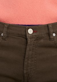 Detailní záběr na hnědé kalhoty zapnuté v pase přes vícerozměrný pletený svetr v růžové a fialové barvě.