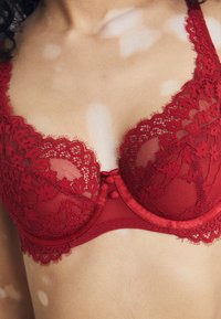 Red lace bra featuring floral patterns, sheer mesh sections, scalloped edges, and decorative buttons at the center front. Elegant design.