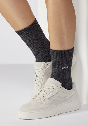 Hellgraue Wildledersneaker mit strukturiertem Obermaterial und weißen Akzenten, kombiniert mit dunkelgrauen gerippten Stricksocken mit weißer Beschriftung.