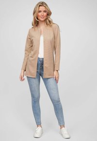 Beige svetr s dlouhým rukávem, rovného střihu a žebrovanou strukturou, nošený přes bílý top a spárovaný s světle modrými skinny džíny a bílými teniskami.