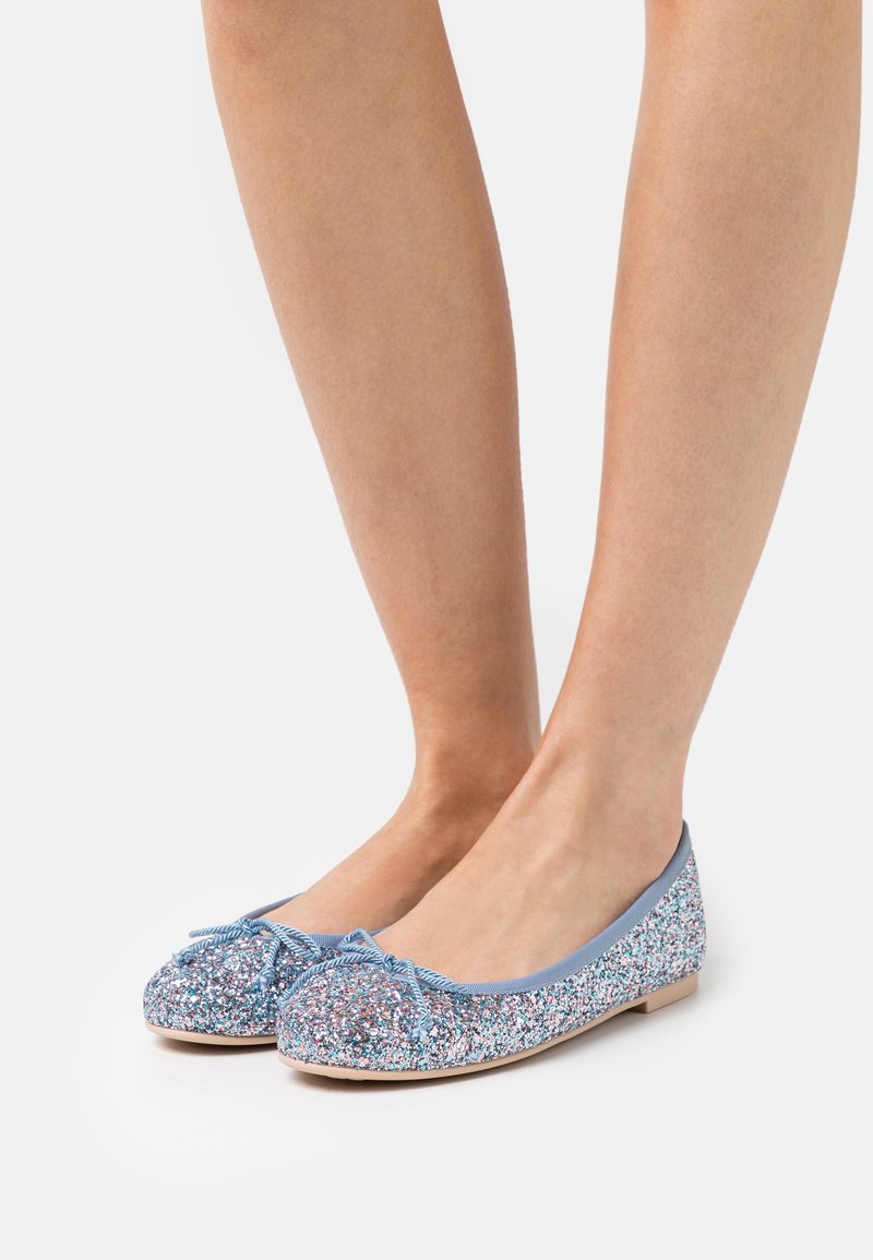 Jambes portant des ballerines bleues et argentées scintillantes avec de petits nœuds, présentées sur un fond blanc uni.