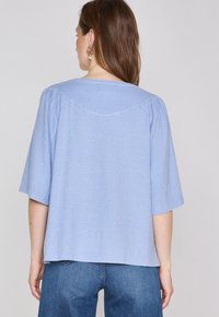 Top azzurro chiaro a maniche corte con dettaglio di spalla arricciata e scollatura arrotondata. Tessuto morbido con una vestibilità rilassata, abbinato a jeans blu.