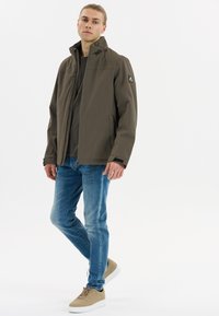 Olivengrøn vandtæt jakke med høj krave, lynlås foran og sidelommer. Båret over en mørk skjorte og parret med blå jeans.