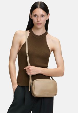 Borsa a tracolla in pelle beige con una superficie liscia, forma arrotondata e tracolla regolabile, tenuta da un modello che indossa un top a costine marrone.