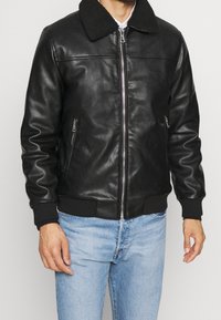 Czarna skórzana kurtka typu bomber z kołnierzem ze sztucznego owczej skóry, zamkiem błyskawicznym z przodu i ściągaczami na rękawach. Posiada boczne kieszenie na suwak oraz gładką teksturę.