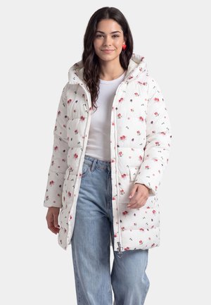 Frau, die eine weiße Daunenjacke mit kleinem roten Blumenmuster, hellblaue Jeans, ein weißes Oberteil und rote herzförmige Ohrringe trägt, lächelt in die Kamera.